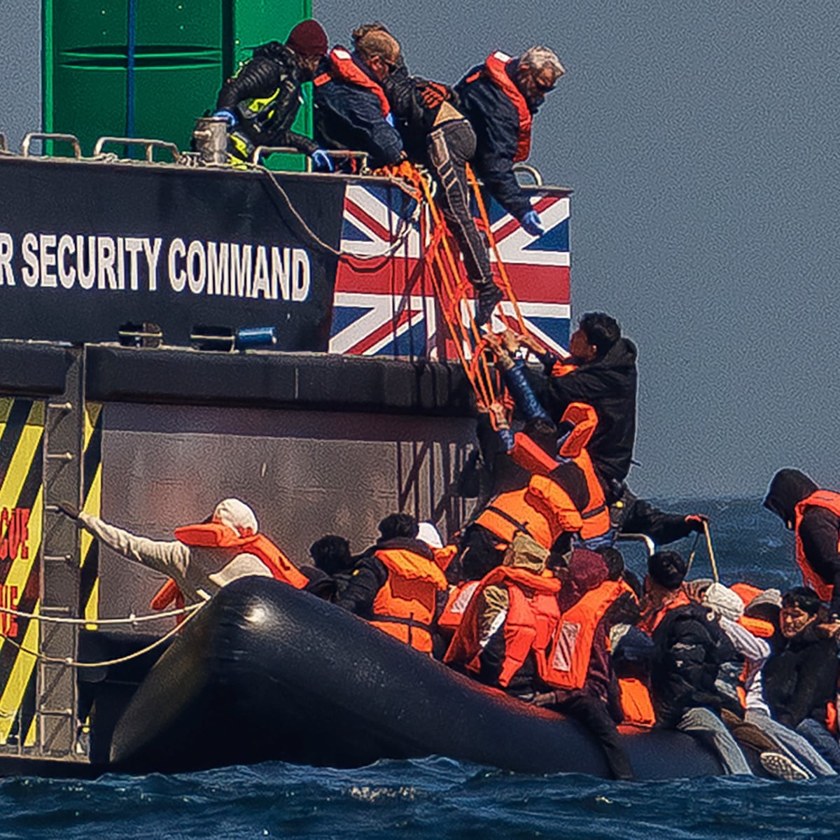 People in orange life vests board a UK Security Command vessel from an inflatable raft at sea. People in orange life vests board a UK Security Command vessel from an inflatable raft at sea.
