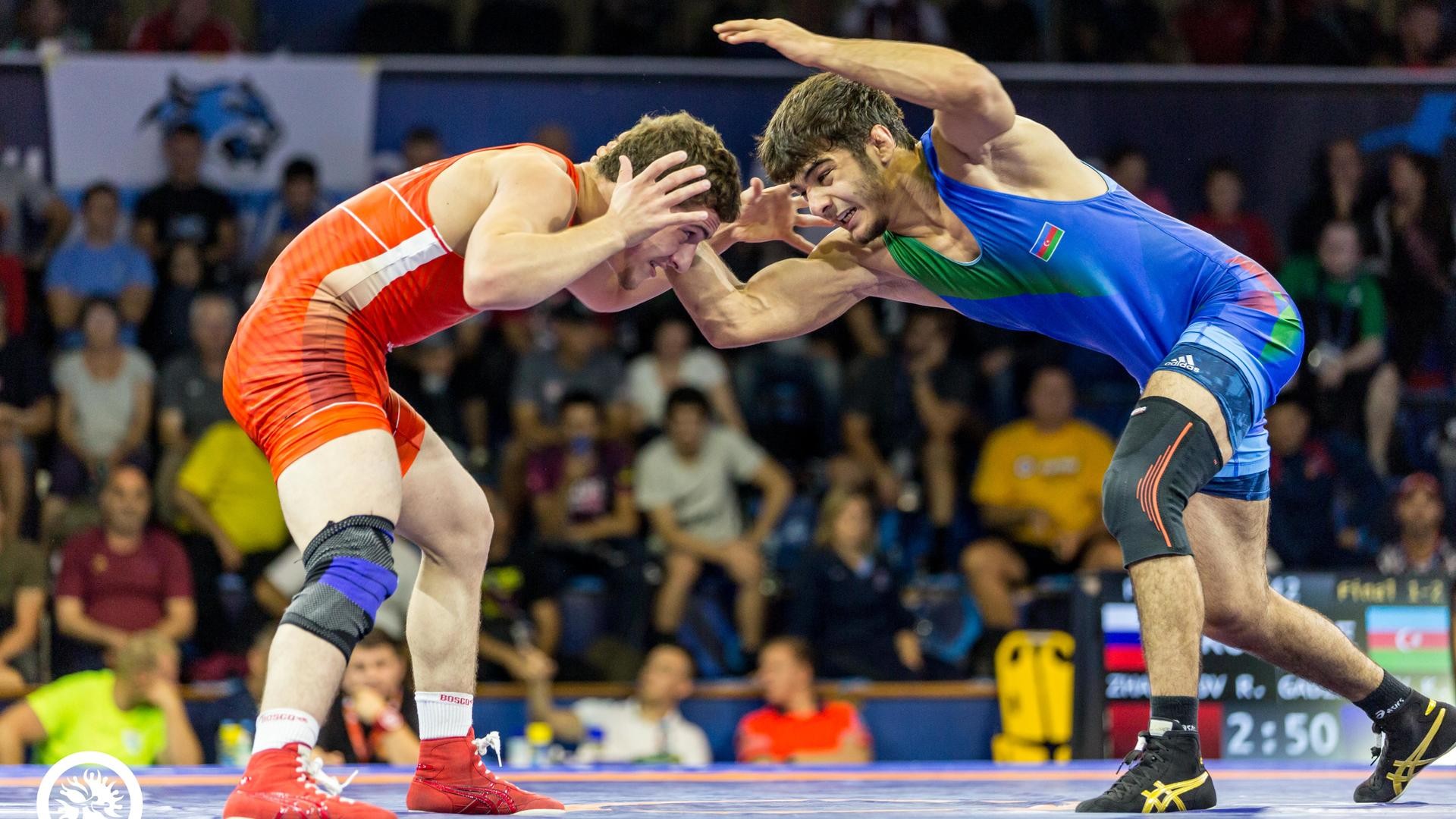 Two male wrestlers grapple on a mat during an indoor sports competition with spectators present. Two male wrestlers grapple on a mat during an indoor sports competition with spectators present.