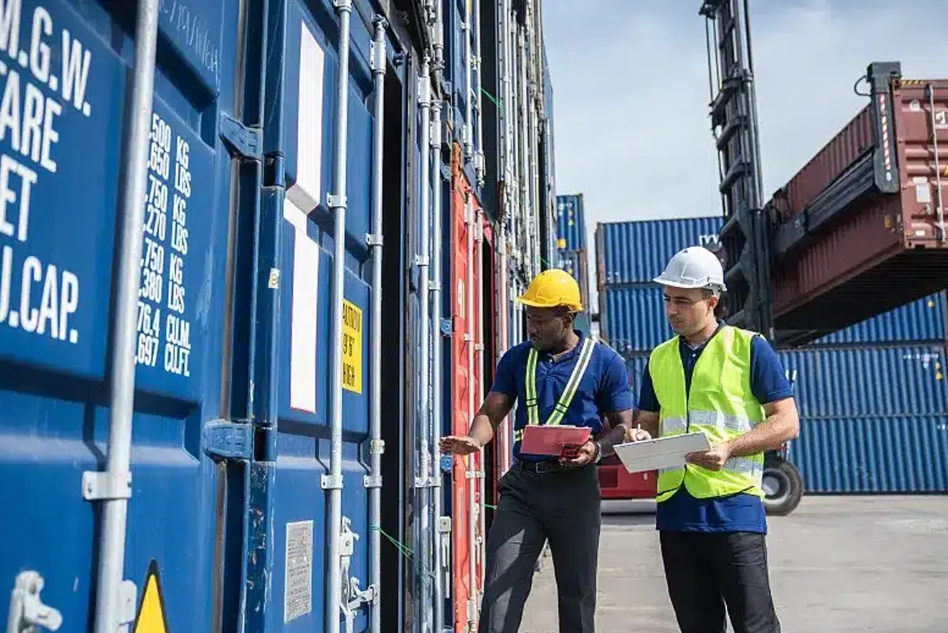 Two workers in hard hats inspect shipping containers at a port facility. Two workers in hard hats inspect shipping containers at a port facility.