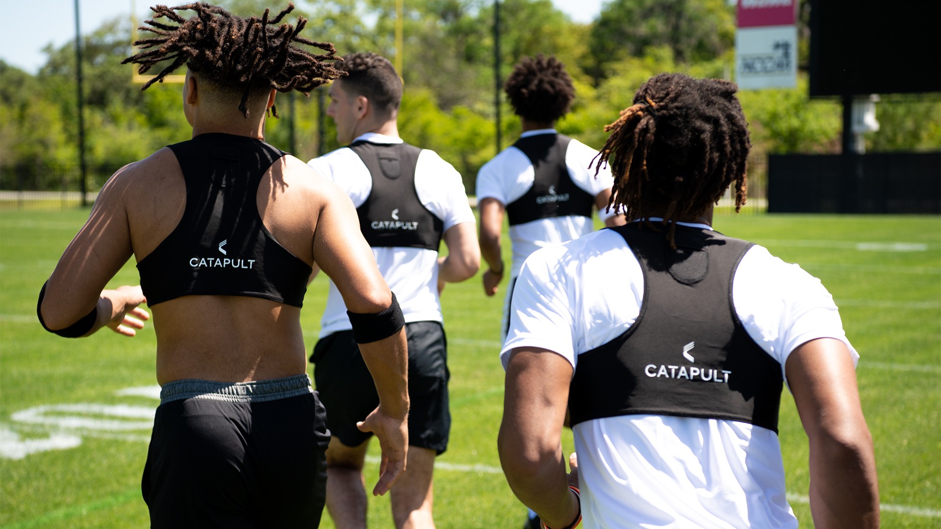 Four male athletes in Catapult vests run across a grassy sports field.