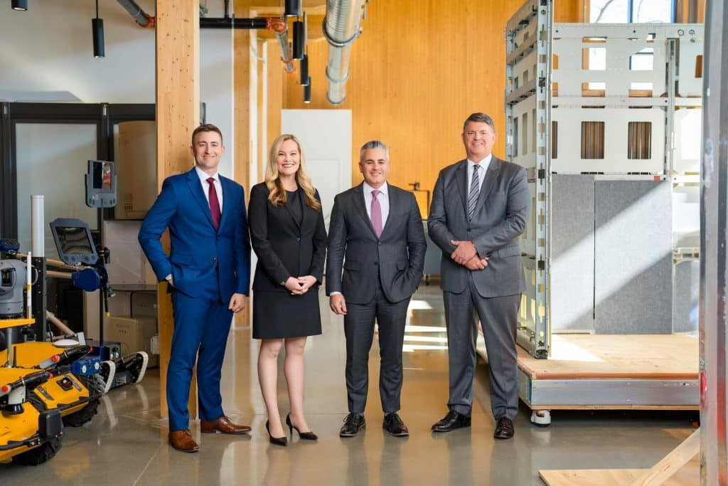 Four professionals in business attire pose together in an industrial facility with machinery visible. Four professionals in business attire pose together in an industrial facility with machinery visible.