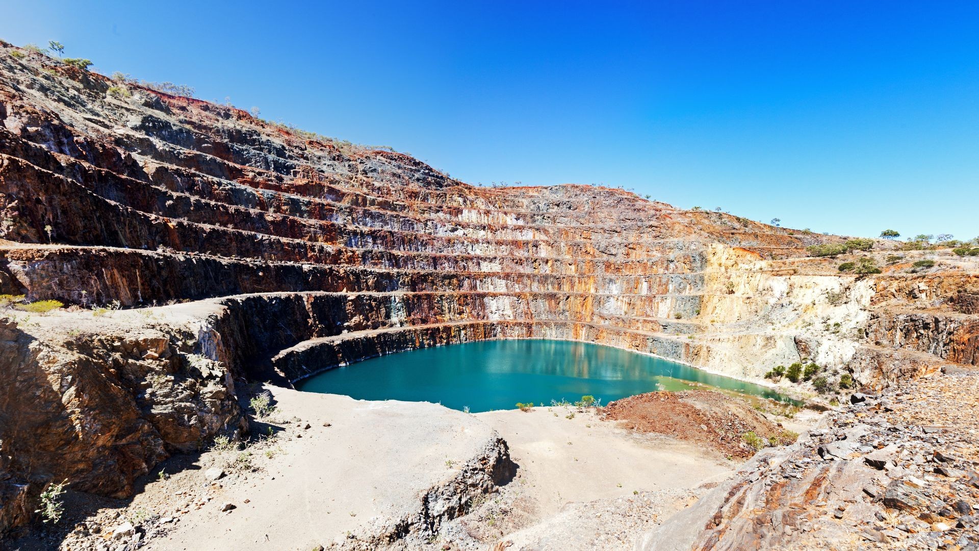 An open-pit mine with terraced rock walls holds a turquoise water pool in an arid landscape. An open-pit mine with terraced rock walls holds a turquoise water pool in an arid landscape.