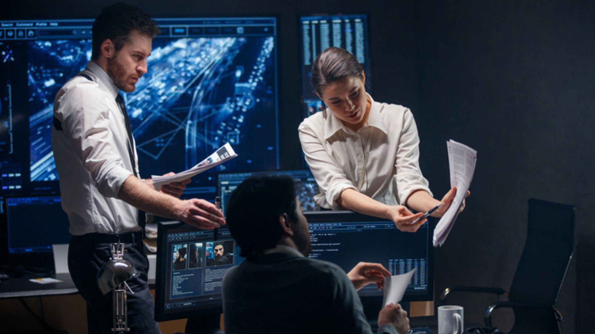 Three professionals review documents and monitor systems in a dimly lit tech control room. Three professionals review documents and monitor systems in a dimly lit tech control room.