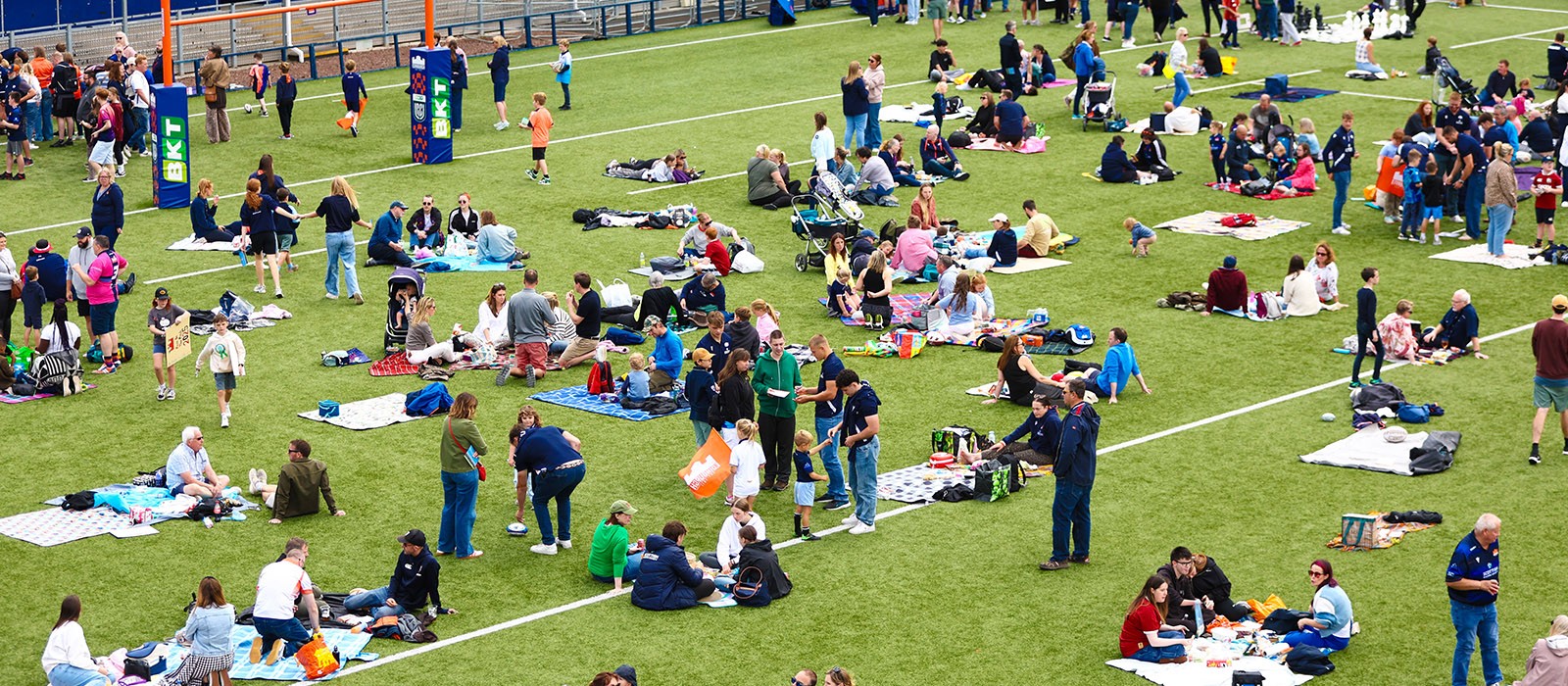 Many people gather on a grassy sports field, sitting on blankets and interacting. Many people gather on a grassy sports field, sitting on blankets and interacting.