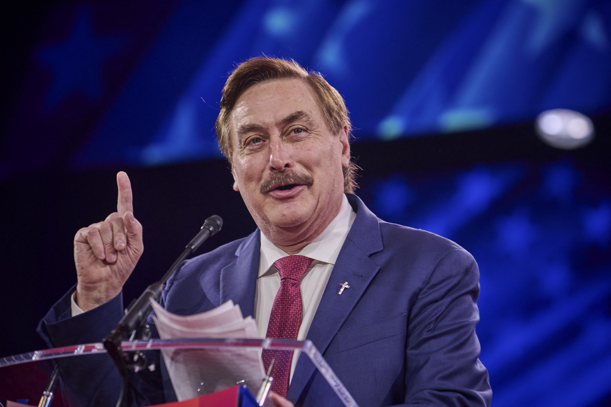 Man in blue suit speaks at podium, pointing finger under blue stage lighting. Man in blue suit speaks at podium, pointing finger under blue stage lighting.