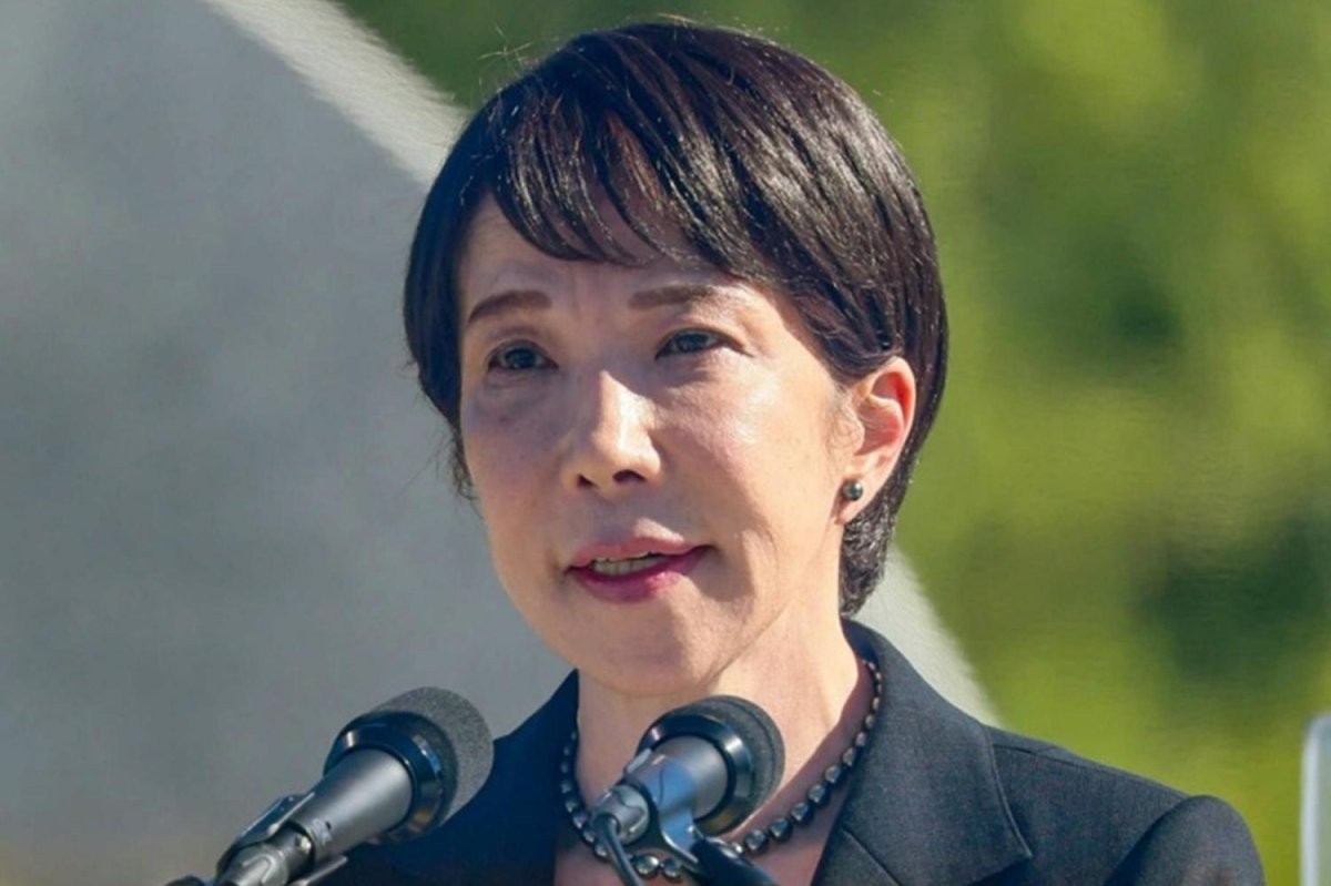 A woman speaks into two microphones outdoors with blurred greenery and a white structure in background.