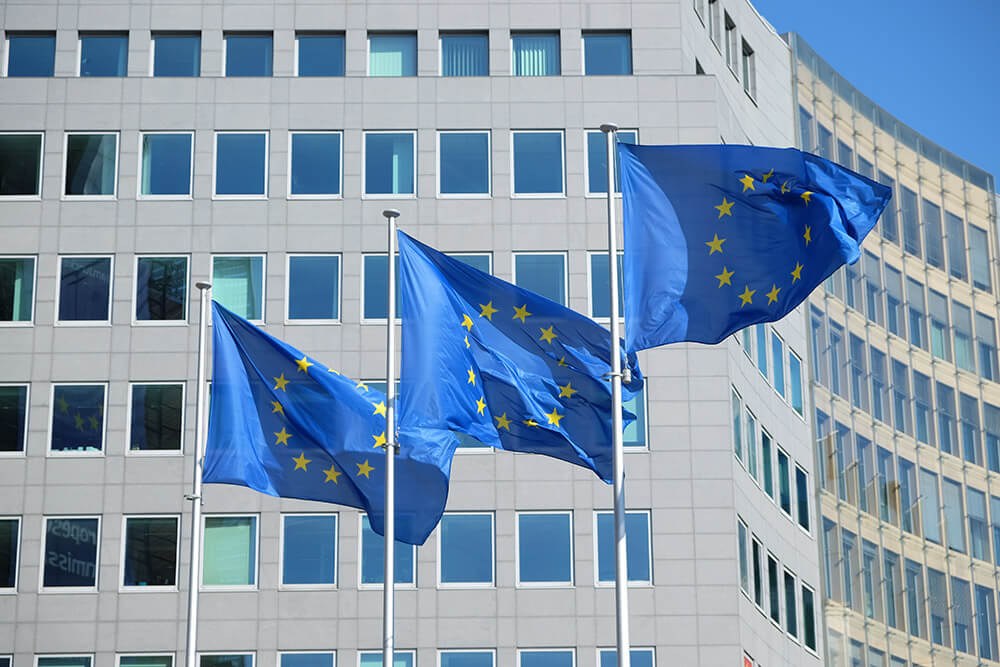 Three European Union flags wave in front of a modern office building with many windows. Three European Union flags wave in front of a modern office building with many windows.