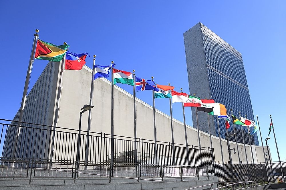 Multiple national flags wave before a large building and adjacent skyscraper under clear blue sky. Multiple national flags wave before a large building and adjacent skyscraper under clear blue sky.