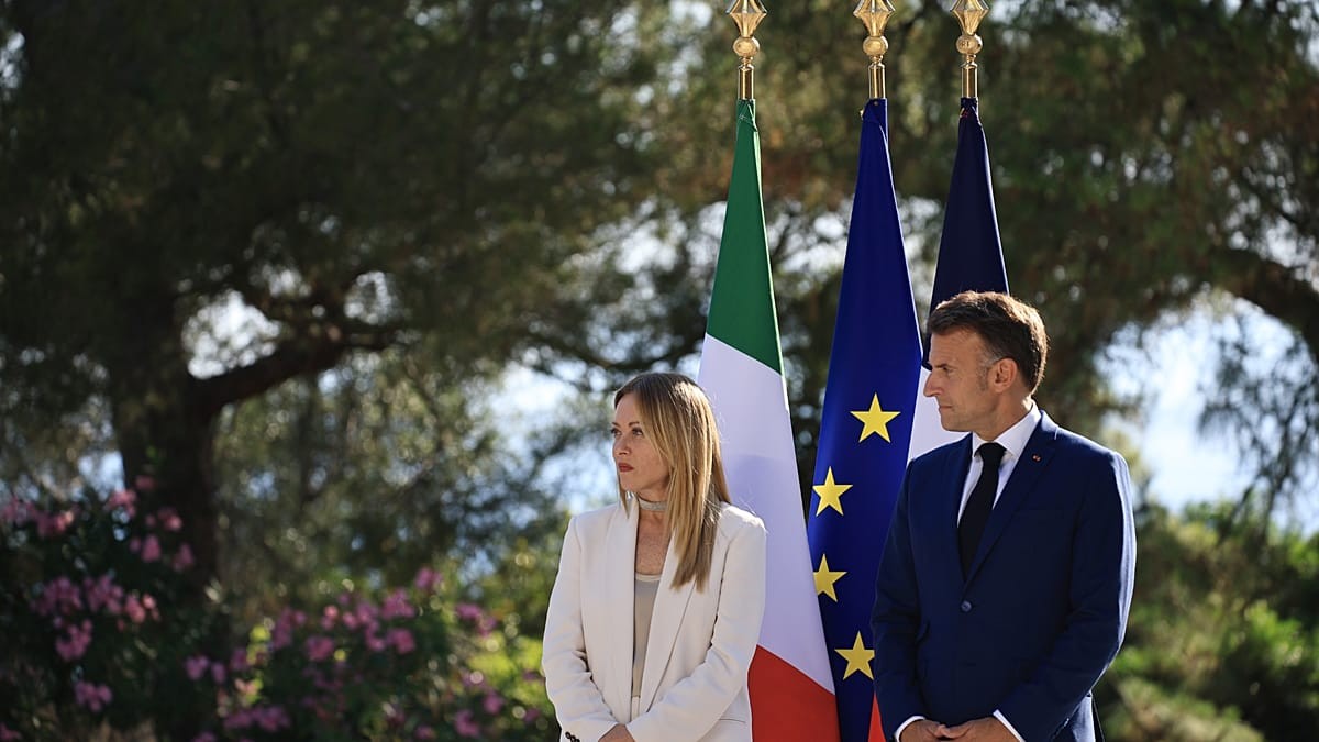 A woman in white suit and a man in blue suit stand before three flags outdoors. A woman in white suit and a man in blue suit stand before three flags outdoors.
