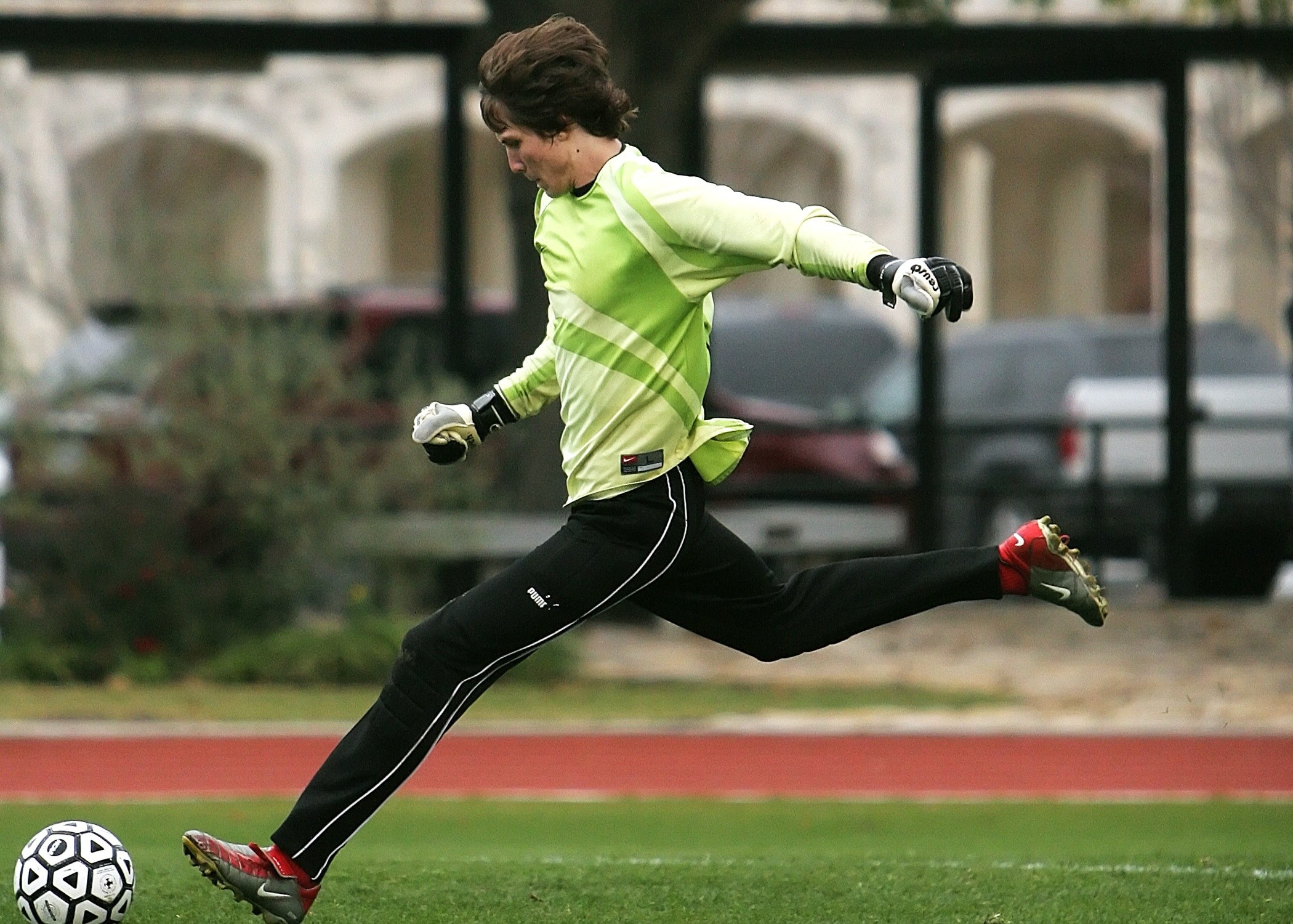 A male soccer goalkeeper kicks a ball on an outdoor grass field near buildings. A male soccer goalkeeper kicks a ball on an outdoor grass field near buildings.