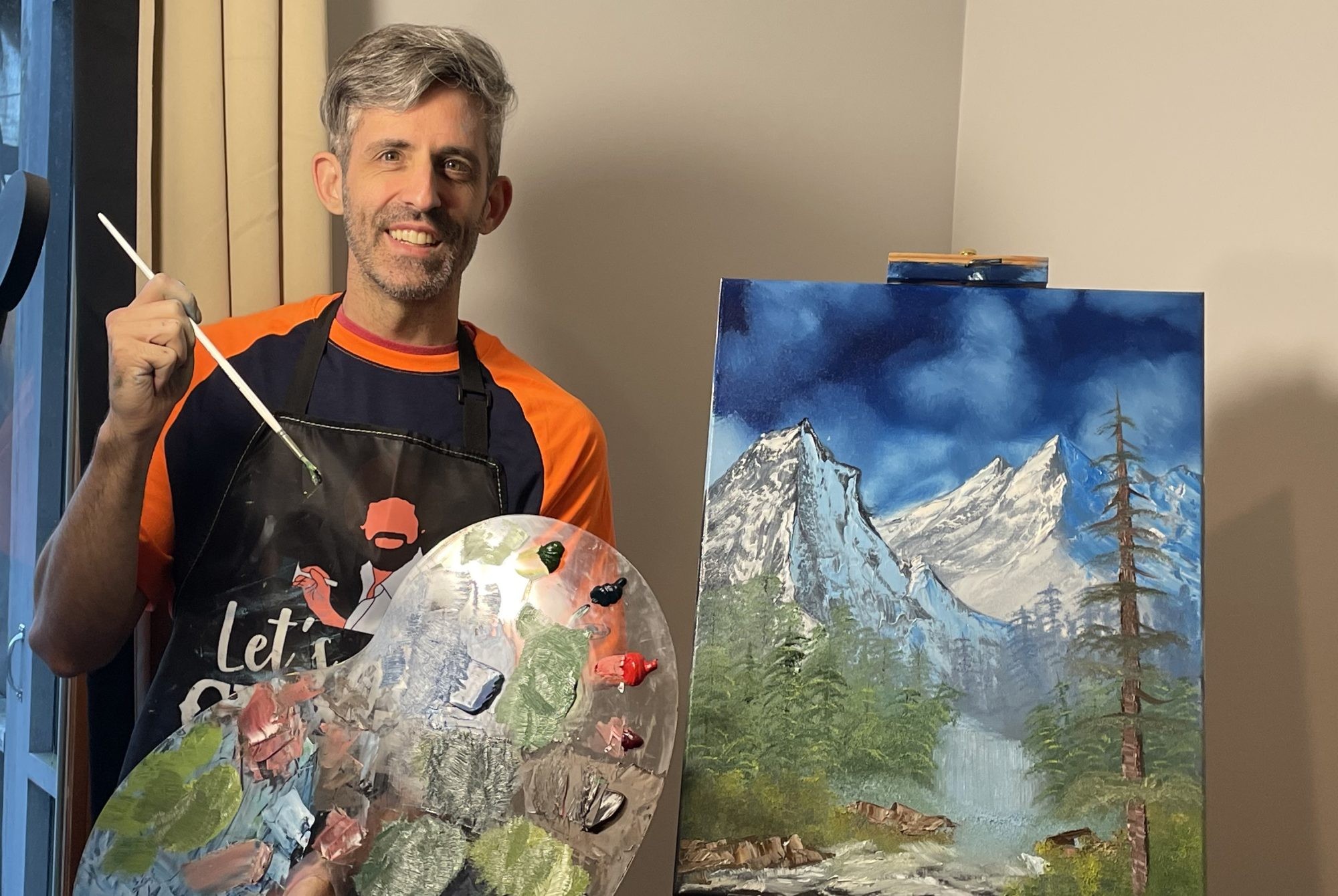 Man holds paintbrush and palette beside a mountain landscape painting on an easel. Man holds paintbrush and palette beside a mountain landscape painting on an easel.